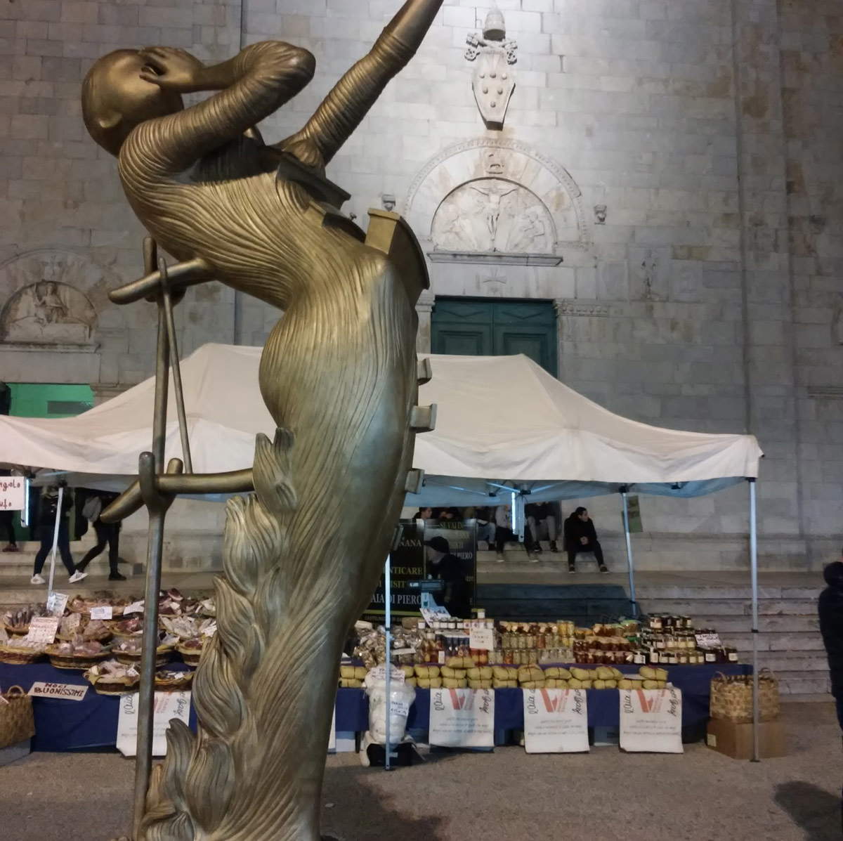 Una delle sculture in piazza del Duomo sullo sfondo di salumi e caciotte