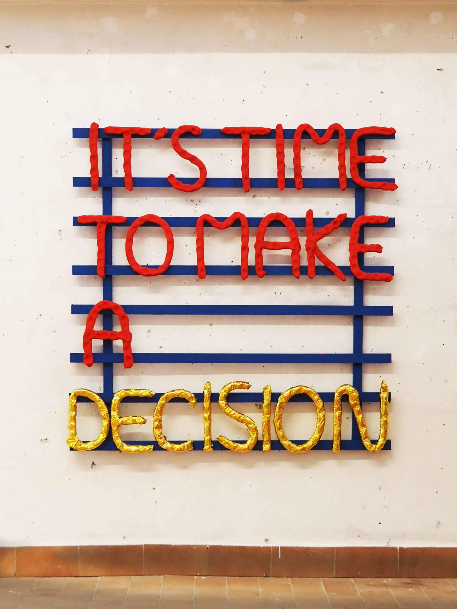 Fabrizio Dusi, It's time to make a decision (2025; Installation aus roter und goldener Keramik, die mit Magneten auf einer blau gestrichenen, mit Nägeln aufgehängten Eisenstruktur angebracht ist, 100 x 100 cm). Mit Genehmigung des Künstlers
