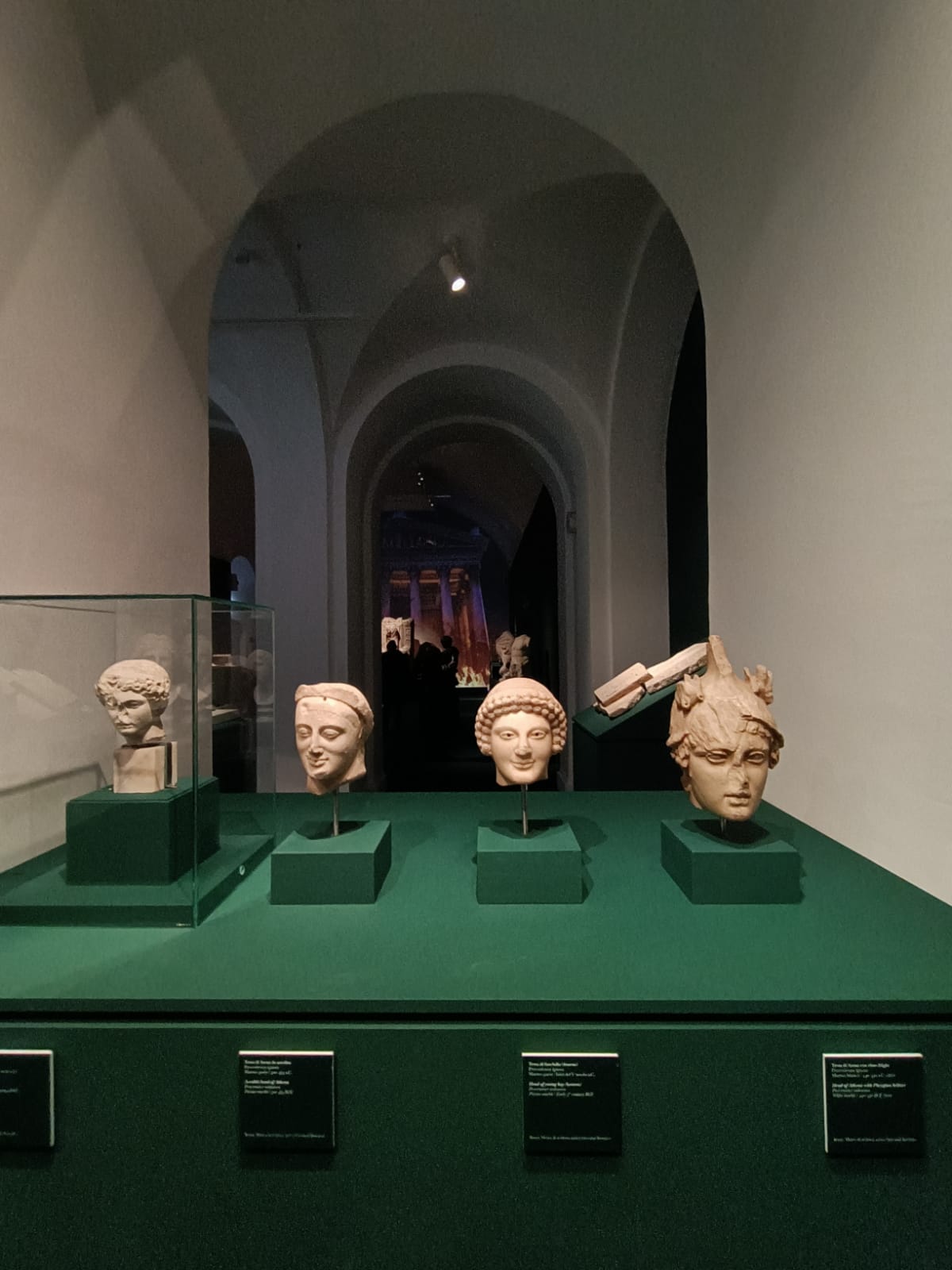 Aufstellung: Die kleine Wand, an der exquisit gearbeitete Köpfe, die zu Kult- oder Votivstatuen gehören, aufgereiht sind, rahmt den großen Raum mit der Videoprojektion ein.